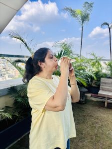 A woman blows into a conch shell. She is wearing a yellow T shirt and standing on a balcony with plants.