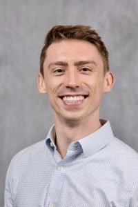 Dr Jacob McCoy is wearing a pale blue shirt and smiling broadly.