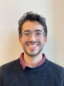 Dr Lopez Servantes has short dark curly hair and stubble. He's wearing a grey sweater and clear-rimmed glasses and he's smiling.