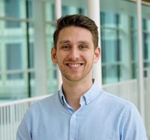 Dr Martinus Oppelaar has short brown hair. He is wearing a light blue shirt standing in front of a modern glass building and smiling.