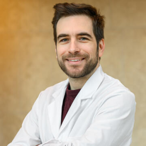 Prof Pengo has short dark hair and beard, he is wearing a lab coat, arms folded and smiling.